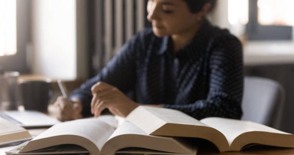 Remote learning. Diligent millennial mixed race female student prepare for exam at home. Confident indian woman write up notes thesis for research report essay. Focus on opened paper books on table